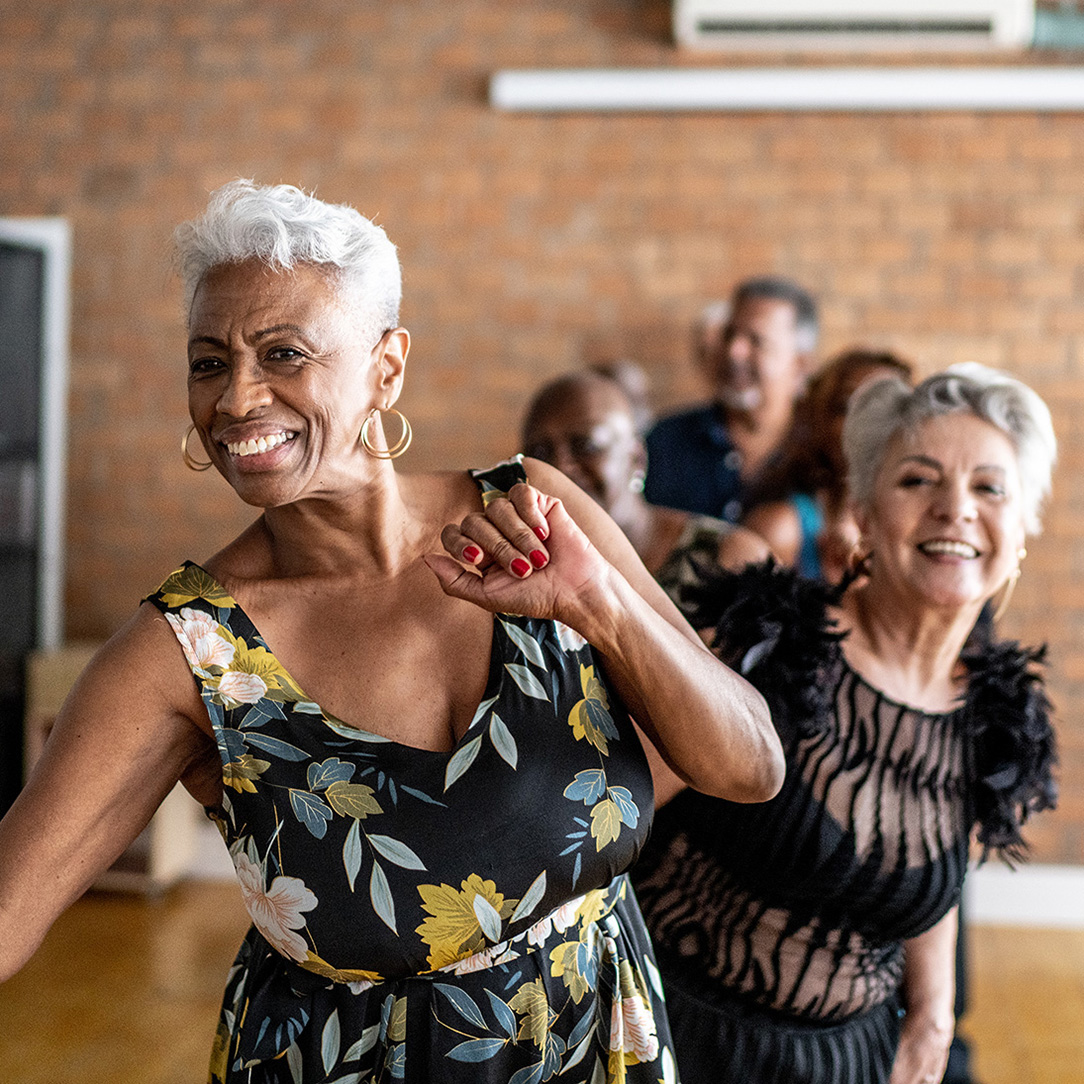 Older adults dancing in a room with a brick wall and wooden floor; one wears a floral dress, another a black dress with feathered sleeves.