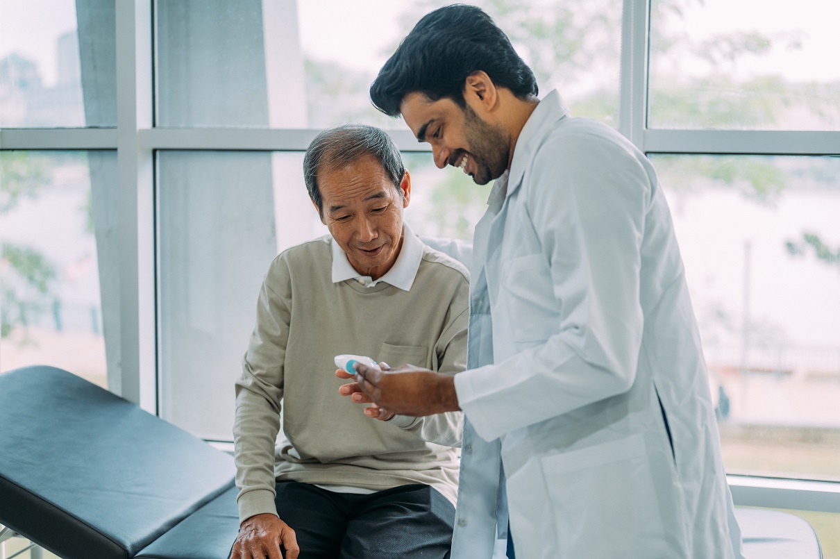 A healthcare professional in a white lab coat stands beside an older adult seated on an exam table, demonstrating how to use a small medical device during a clinical visit. Large windows in the background let in natural light, creating a calm medical setting.
