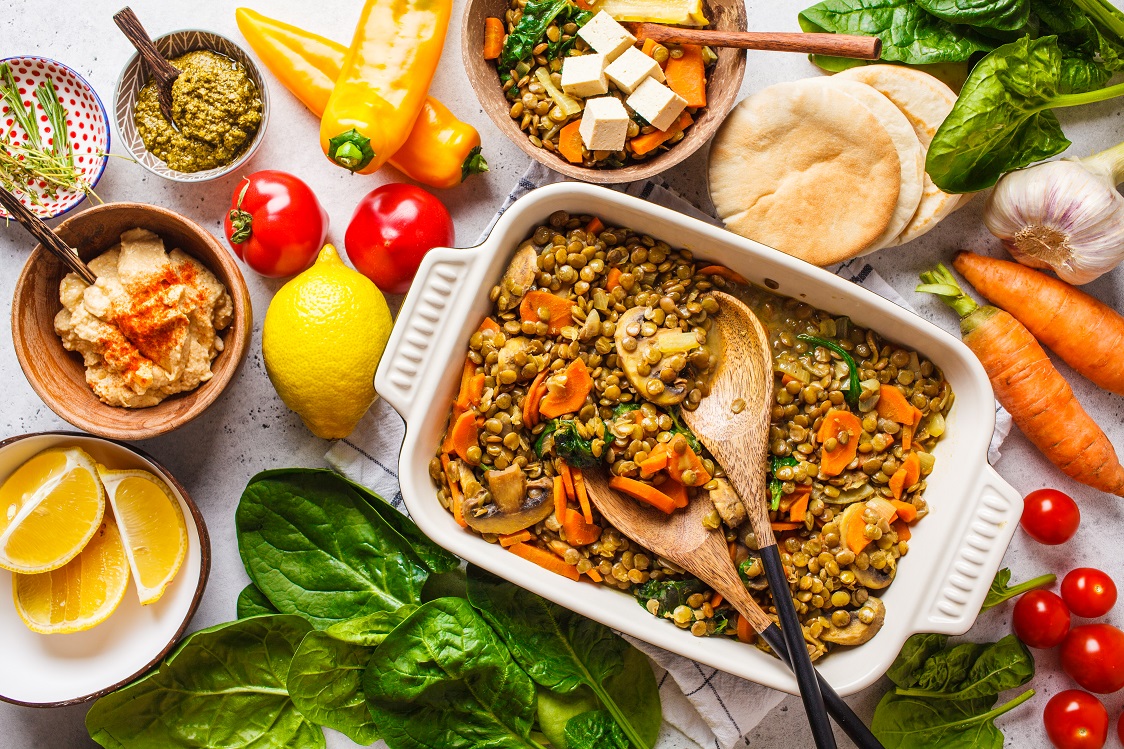 A colorful spread of fresh ingredients and dishes, including a large baking dish filled with lentils and vegetables, surrounded by hummus, lemon wedges, pita bread, spinach, peppers, tomatoes, carrots, garlic, and small bowls of sauces and seasonings.