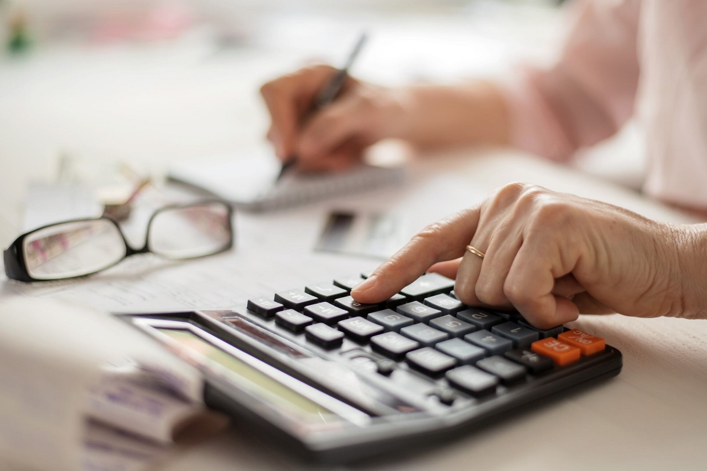 A person uses a calculator while writing notes on paper, with eyeglasses and documents placed on the desk nearby.