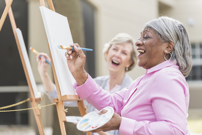 Two people paint on canvases set up on easels outdoors, with one person in the foreground holding a palette and brush while adding color to their canvas.